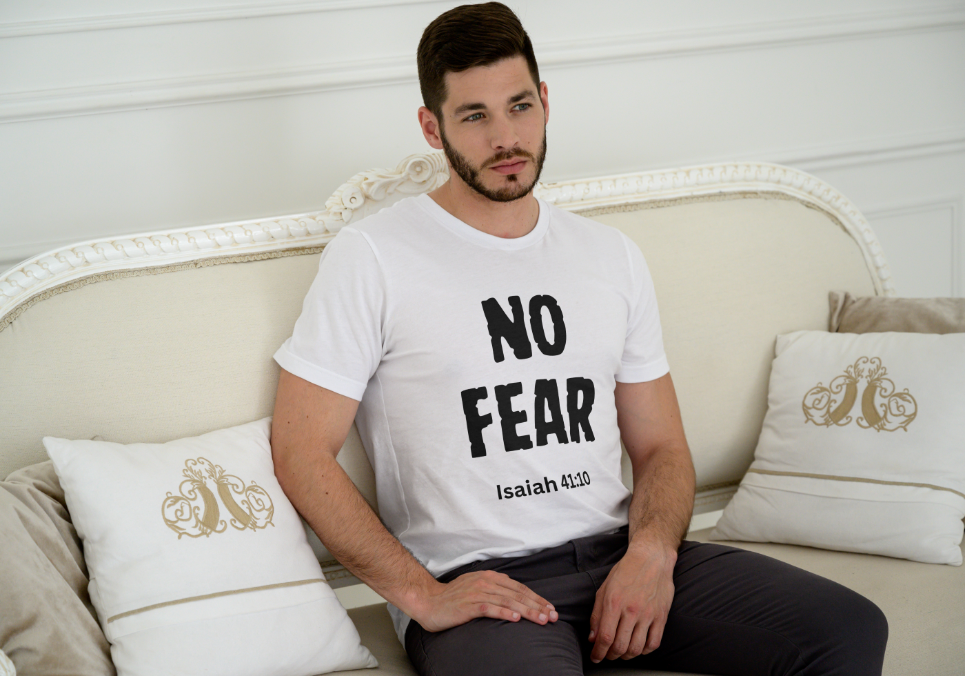 Man sitting on a couch wearing a white t-shirt with 'NO FEAR' text, sitting in a room with decorative pillows.